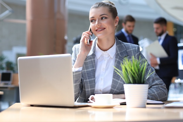 オフィスのコンピューターの近くでスマートフォンを使用して若い笑顔のビジネス女性。