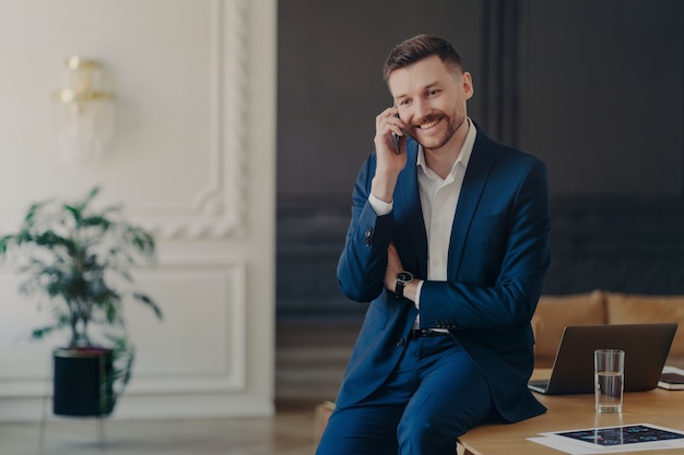 Young smiling businessman in formal dark blue suit talking on phone in stylish office being happy to hear good news while sitting on work desk with laptop, project photos and glass of water