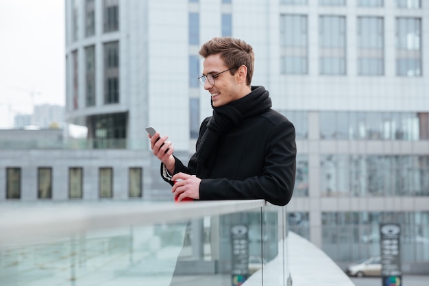 Young smiling Businessman in glasses looking at phone and holding coffee in hand outdoors