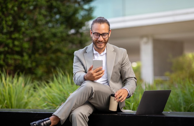 Young smiling businessman using smartphone and drinking takeaway coffee outdoors