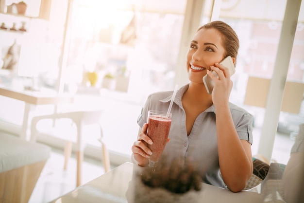 カフェで休憩中の若い笑顔の実業家。彼女はスマートフォンを使ってフルーツスムージーを飲んでいます。