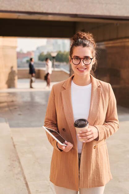 Молодая улыбающаяся деловая женщина в очках и формальной одежде с бокалом кофе на фоне современного здания в городской среде