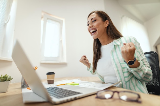 Giovane imprenditrice sorridente sta lavorando al computer in ufficio. sta guardando qualcosa sul laptop e festeggia il successo.