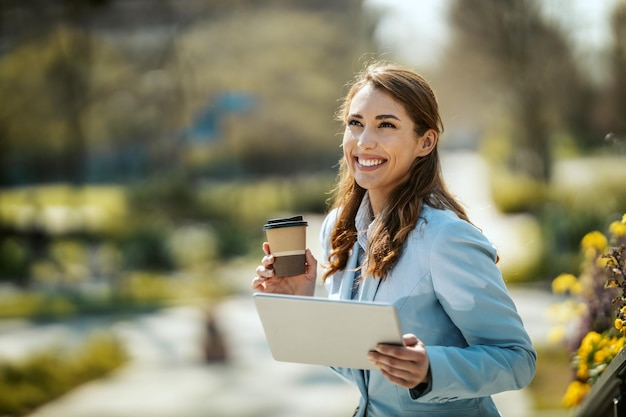 彼女の手にタブレットを持つ若い笑顔の実業家は、コーヒーブレイク中にオフィスの前に立っています。