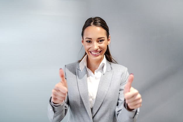 Young smiling caucasian businesswoman in formal wear standing inside corporate firm and showing thumbs up.