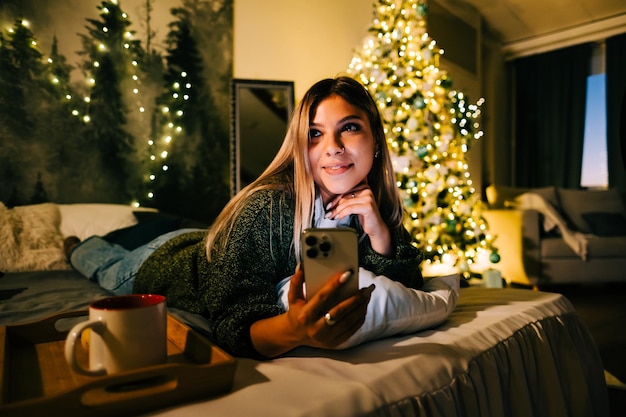 사진 휴일에 집에서 침대에서 휴대전화를 사용하여 웃고 있는 젊은 백인 여성.