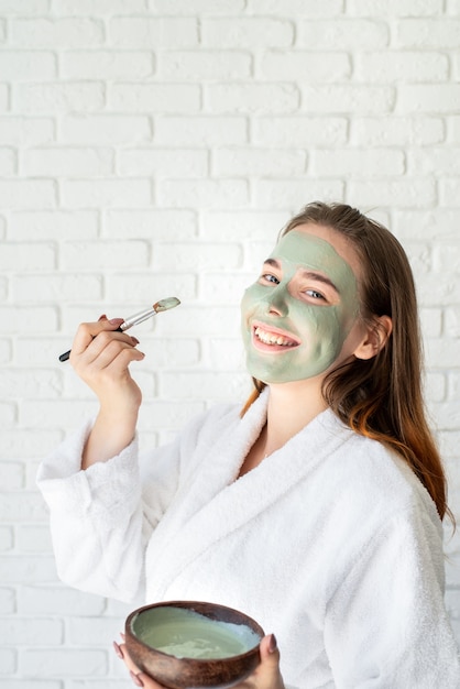 Young smiling caucasian woman wearing bathrobes appplying clay face mask making funny face