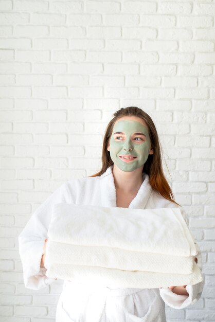 Young smiling caucasian woman with a face mask wearing bathrobes holding a pile of white bath towels