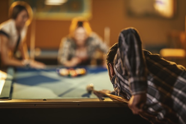 Young smiling cheerful friends are playing billiards in bar after work.  They are involved in recreational activity.