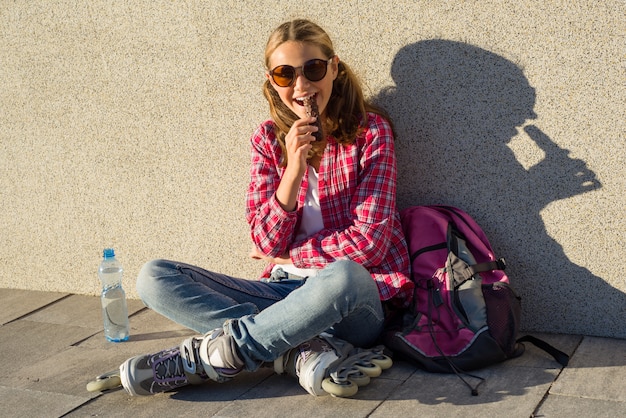 若い笑顔のクールな女の子、チョコレートバーを食べるローラースケートで履いた