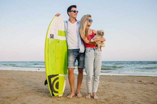 Young smiling couple having fun on beach posing with surf board playing with dog