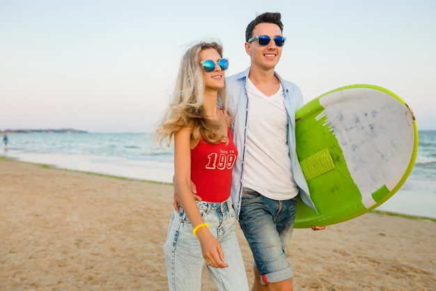 Young smiling couple having fun on beach walking with surf board