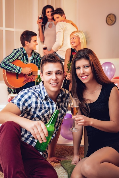 Young smiling couple at home party. They are sitting on the floor with drinks and looking at camera. In the background their friends talking and playing acoustic guitar.