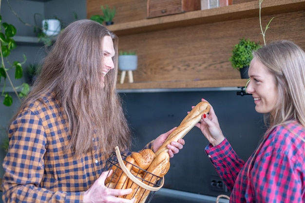 A young smiling couple in plaid shirts take a French baguette from a basket in a cozy kitchen