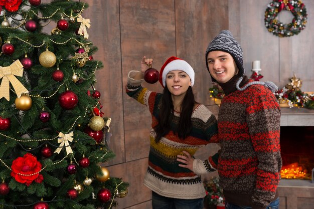 Young smiling couple wearing funny hats, decorating for Christmas hanging the decorations and ornaments on the large Christmas tree in their living room