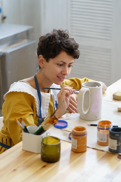 Молодая улыбающаяся творческая женщина, работающая в собственной уютной гончарной мастерской, изготавливающая орнамент на керамическом кувшине