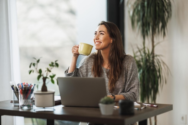 Young smiling designer woman holding a cup of coffee and using a laptop while working from home.