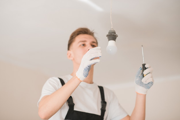 Young smiling electrician with a screwdriver repairing a ceiling lamp indoors. Space for text