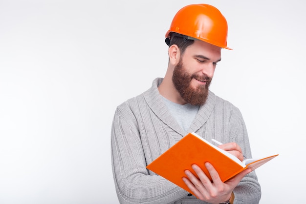 Il giovane ingegnere sorridente che porta il cappello duro sta scrivendo qualcosa nel suo quaderno sulla parete bianca