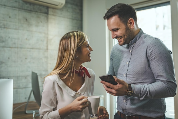 Giovani imprenditori sorridenti stanno facendo una pausa caffè, parlando e usando lo smartphone in ufficio.