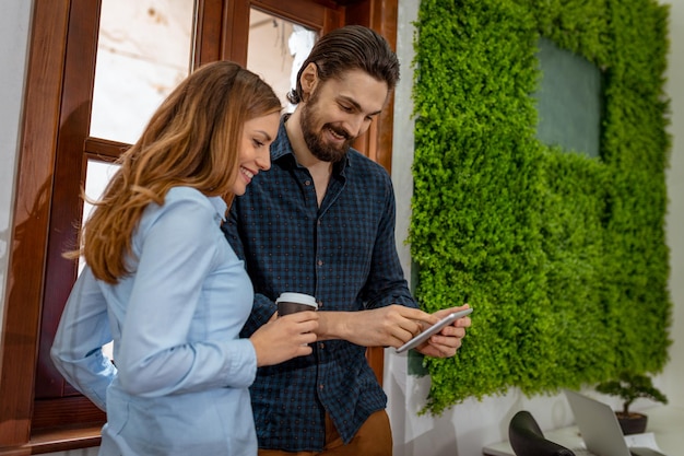 Foto giovani imprenditori sorridenti che fanno una pausa caffè e utilizzano la tavoletta digitale davanti alla finestra dell'ufficio.