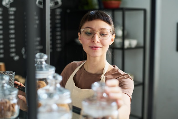 Young smiling female clerk checking herbal tea in small jars