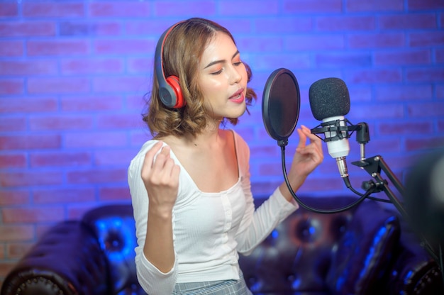 カラフルなライトのある音楽スタジオで曲を録音しながら、マイク付きのヘッドフォンを身に着けている若い笑顔の女性歌手。