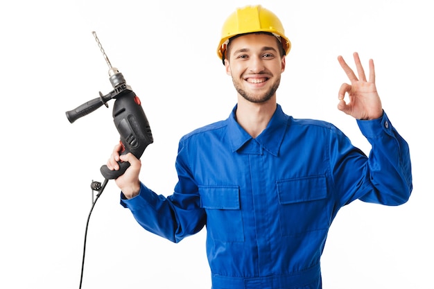 Young smiling foreman in blue uniform and yellow helmet joyfully showing ok gesture lwhile ooking in camera holding drilling machine in hand 