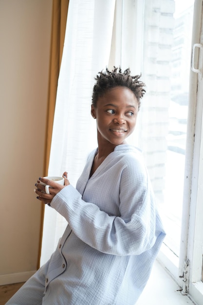 Photo young smiling girl in a beautiful cozy gently blue muslin pajamas stands by the window and thinks