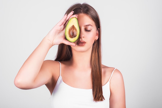 Young smiling girl of European appearance is holding avocado in front of her eye on white background
