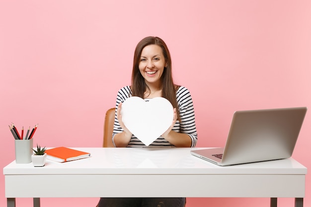 Молодая улыбающаяся девушка держит белое сердце с копией пространства, работая над проектом, сидя в офисе с ноутбуком