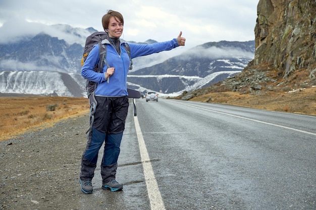 A young smiling girl is traveling in the mountains stops the car on the road hitchhiking raises his hand