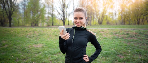 写真 スポーツをしている若い笑顔の女の子は、画面を見ている街で日没時にワイヤレスヘッドフォンで音楽を聞くために彼女の携帯電話を使用して公園で走っています