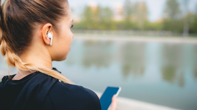 Young smiling girl making sport and running in the park using her phone to listen the music with wireless headphones on sunset in the city watching the screen