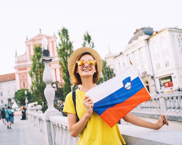 Young smiling girl in sunglasses with slovenian flag on central square of Ljubljana Woman tourist holding slovenian flag on background of city architecture Travel living study in Slovenia Europe