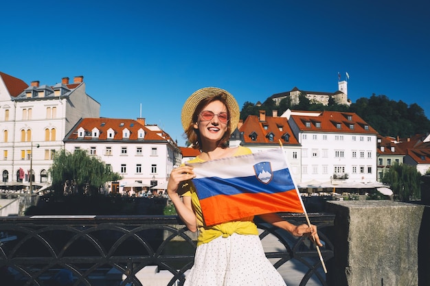 Young smiling girl in sunglasses with slovenian flag on central square of Ljubljana Woman tourist holding slovenian flag on background of city architecture Travel living study in Slovenia Europe