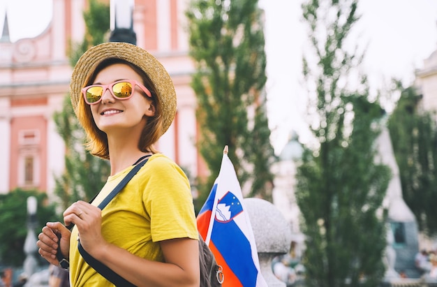 Young smiling girl in sunglasses with slovenian flag on central\
square of ljubljana woman tourist and slovenian flag on background\
of city architecture travel living study in slovenia europe