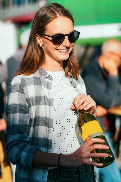 Young smiling girl wearing sunglasses holds wine bottle. Woman casual style. Female person having fun. Happy holiday celebration. Outdoors lifestyle portrait. Adult beautiful face. Street event
