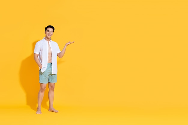 Young smiling handsome Asian man in beach attire doing open palm gesture showing empty copyspace in summer yellow isolated wall