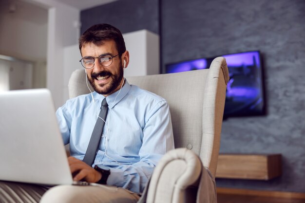 Young smiling handsome bearded businessman sitting in chair at home and using laptop.