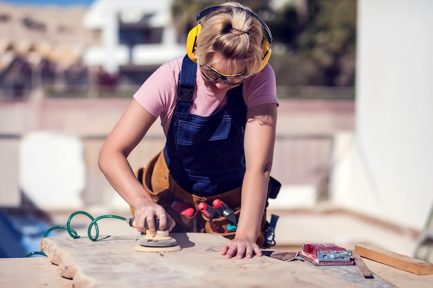 Young smiling handy woman with short blond hair sanding wooden surface.