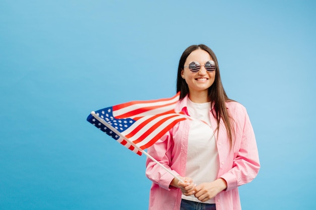 Young smiling happy woman wears casual clothes holding american flag and looking to the camera isolated on blue color background