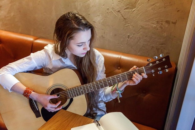 Young smiling hipster woman sitting playing guitar at cafe. Girl learning to play song