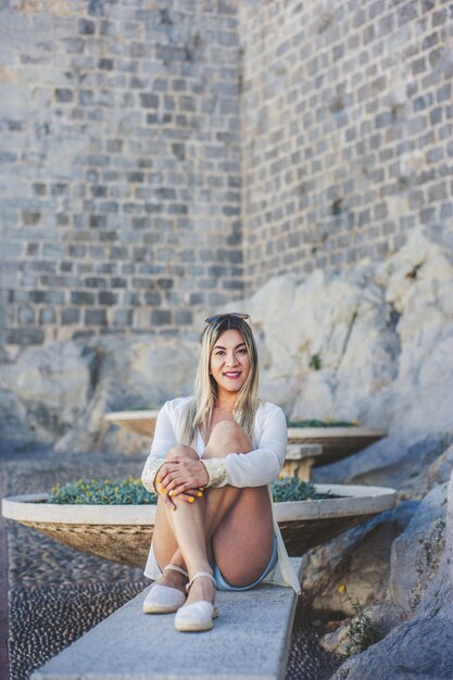 Young smiling Hispanic female sitting on a bench onn old wall under the sunlight