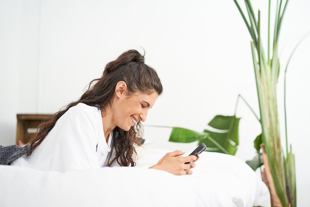 Young smiling hispanic woman uses smart phone lying on bed people using social networks and chatting...