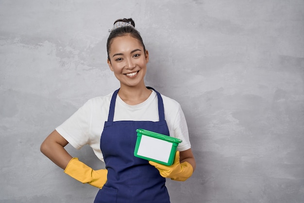 Photo young smiling housewife or maid woman in uniform and yellow rubber gloves holding green plastic