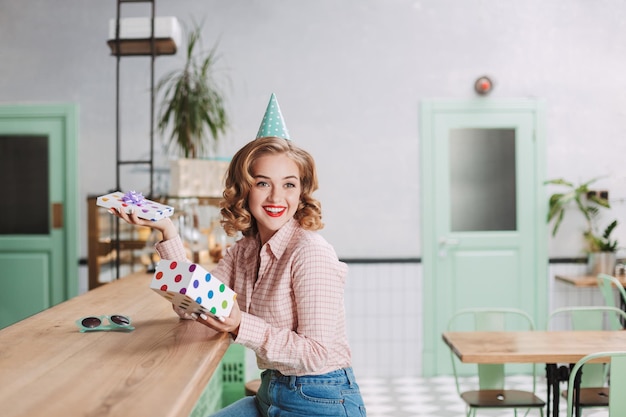 Young smiling lady in birthday cap sitting at the bar counter with open present box in hands and happily looking aside in cafe