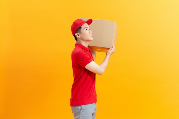 Young smiling logistic delivery man in red uniform holding the box on yellow background