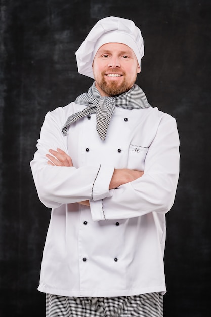 Young smiling male chef crossing arms by chest while looking at you against black background