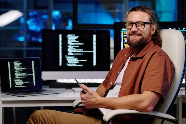 Young smiling male IT engineer in eyeglasses using smartphone